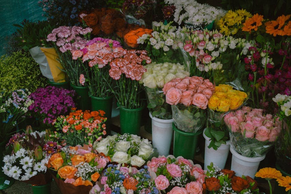 A vibrant display of fresh flowers arranged in green and white vases and buckets, featuring pink roses, yellow roses, white roses, orange carnations, pink eustomas, white daisies, purple chrysanthemum