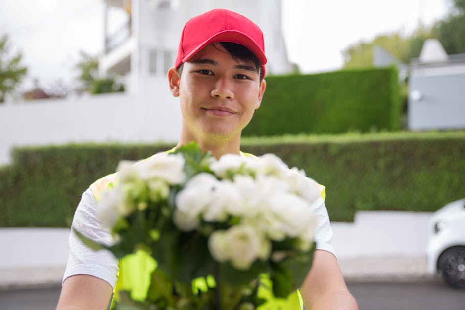 A delivery person wearing a red cap and a high-visibility vest is smiling and holding out a bouquet of fresh white roses and green foliage towards the camera, standing outdoors on a suburban street wi
