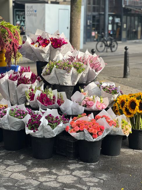 A display of numerous flower bouquets arranged in black plastic pots, set outdoors on a paved area. The bouquets feature a variety of fresh flowers, including pink, purple, white, and red blossoms, wr