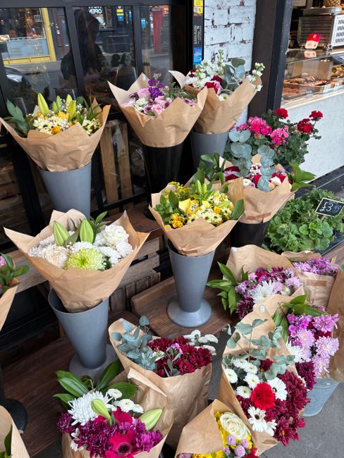 A display of various floral bouquets arranged in tall, sleek, gray vases and wrapped in brown kraft paper on a wooden surface outside a flower shop. The bouquets feature a mix of fresh flowers includi