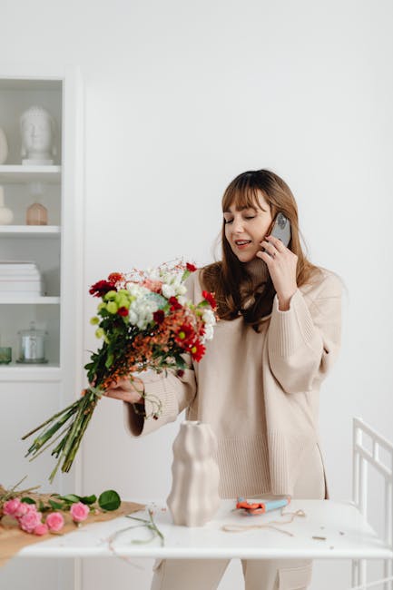 A woman with light brown hair and bangs, wearing a beige sweater, is holding a large bouquet of fresh flowers with a mix of red, pink, white, and green blooms, including roses and chrysanthemums, whil