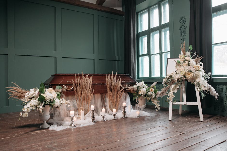 A floral arrangement featuring predominantly white flowers, including roses and lilies, with accents of soft beige dried grasses, arranged on a white stand and surrounding a white urn on the floor. Th