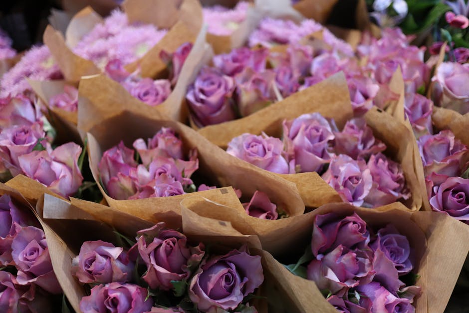 A collection of freshly cut pink and lavender roses arranged in small bouquets wrapped in brown craft paper, positioned closely together on a floral display. The roses have vibrant, velvety petals wit